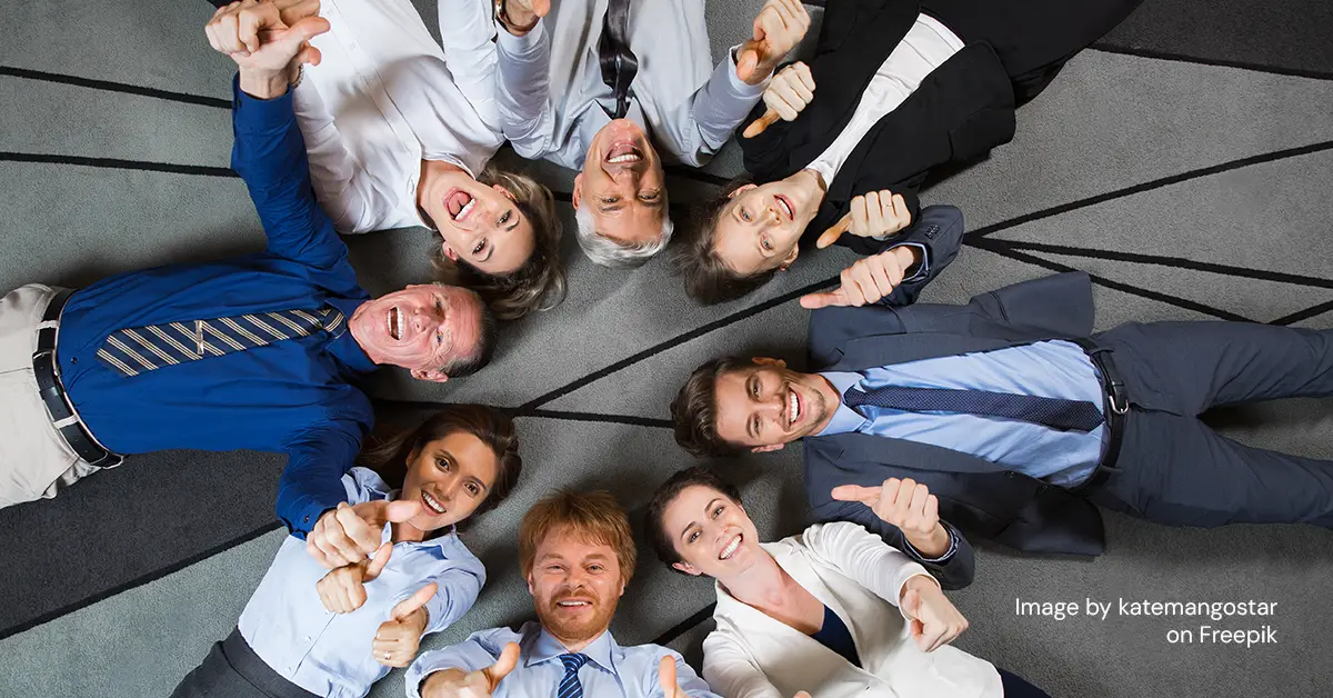 business professionals looking up at camera while forming a circle on the floor and holding two thumbs up