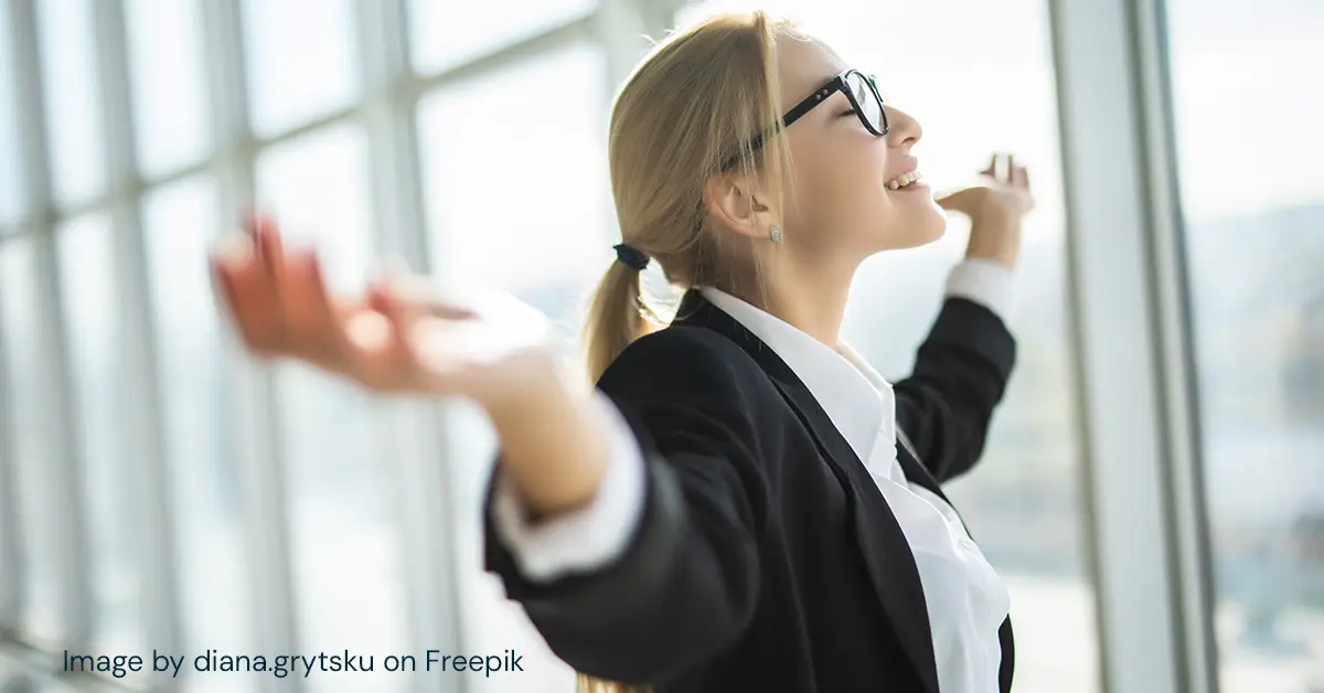 Smiling business woman stands in front of windows with closed eyes and outstretched arms